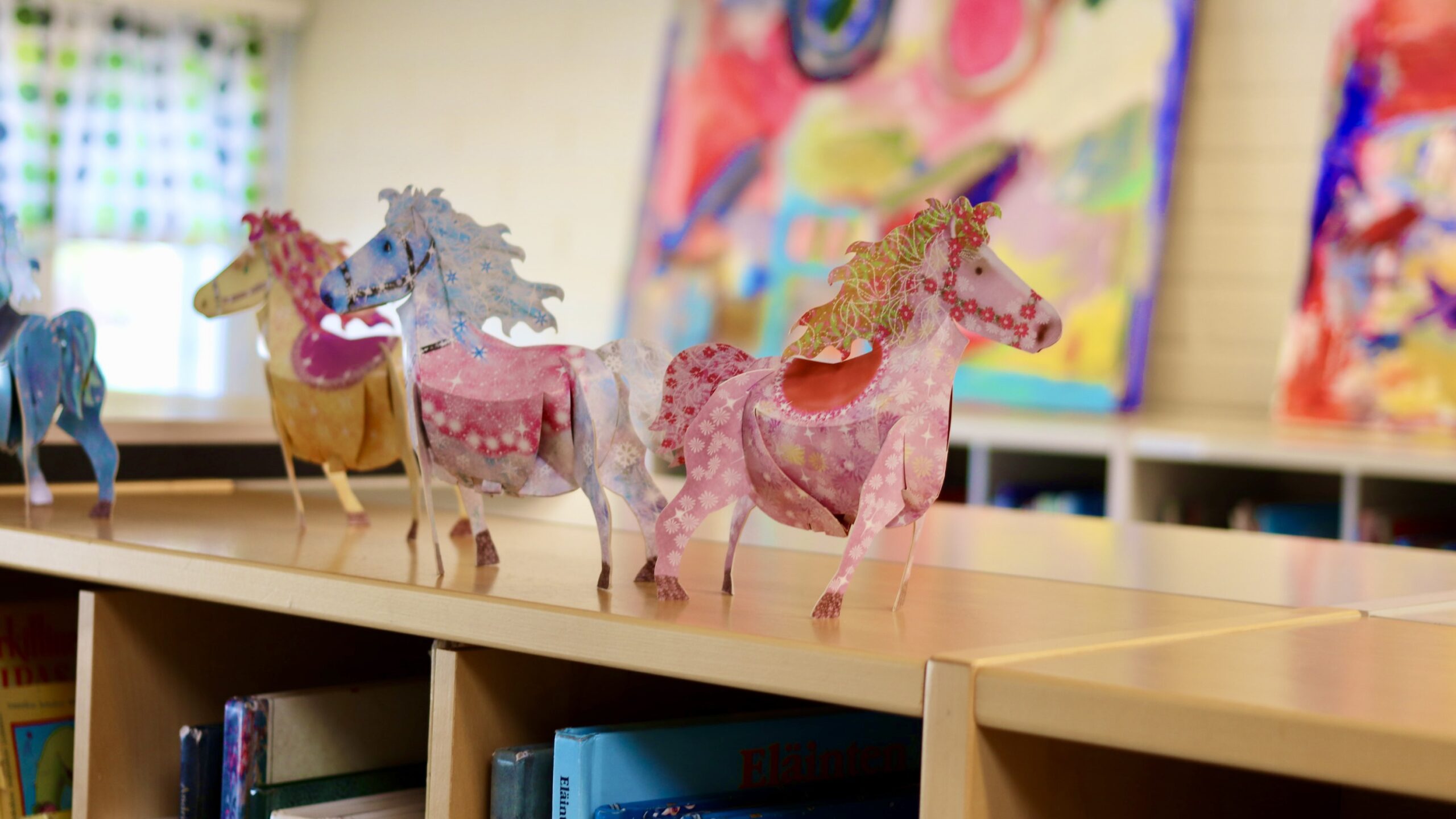 Kolme paperihevosta kirjahyllyn päällä.