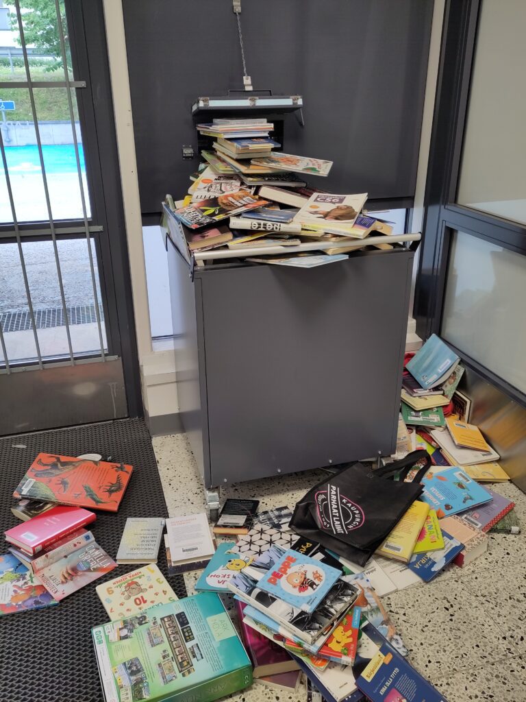 Palautuslaatikko täynnä kirjoja, joista osa pudonnut lattialle.