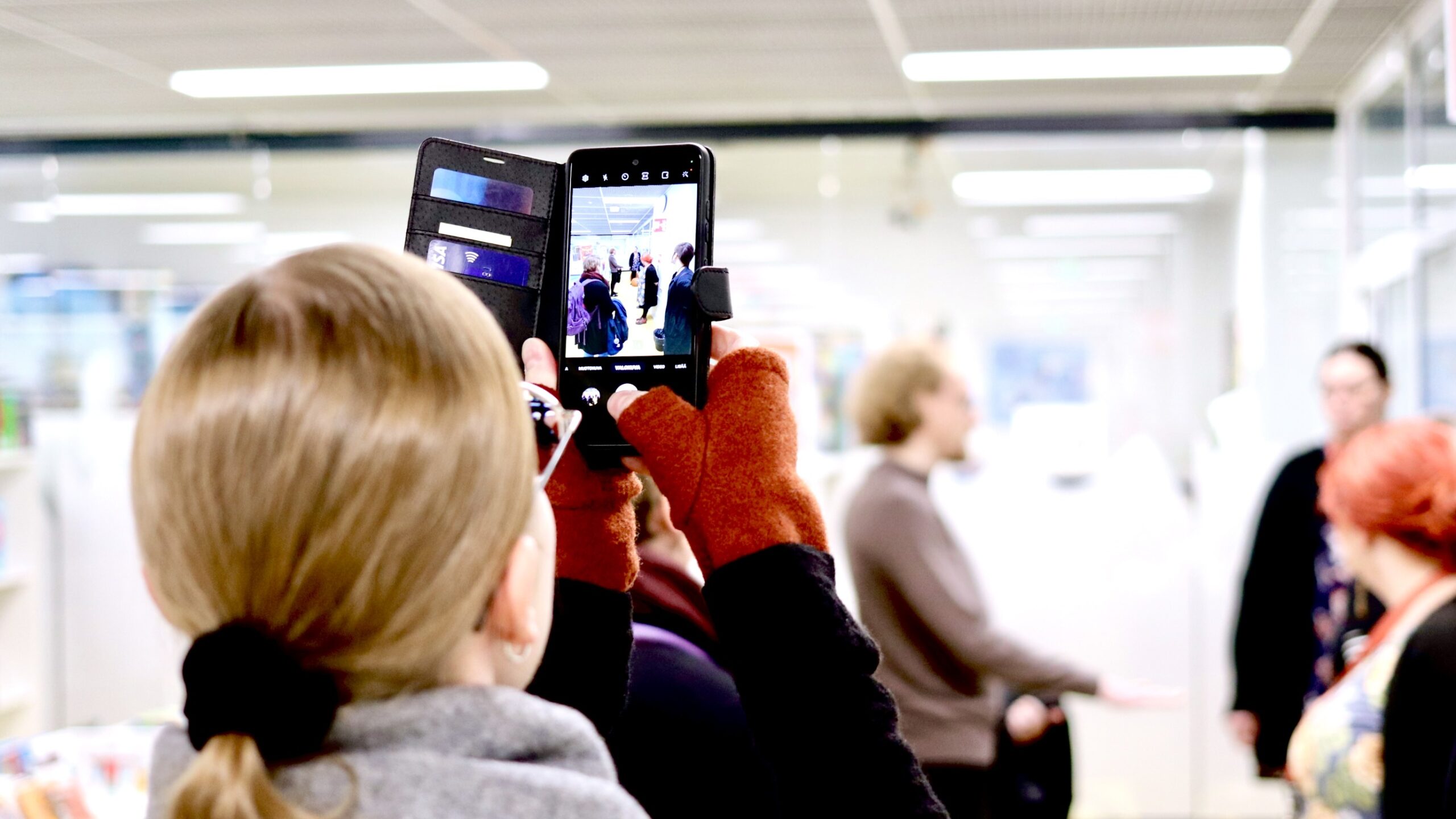 Henkilön pää selin kameraan, kädessä kirjastotilaa ja taustan ihmisiä kuvaava älypuhelin.