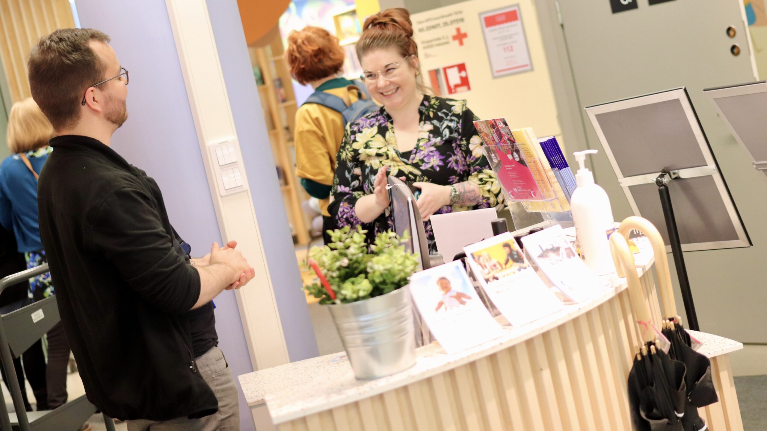 Kaksi ihmistä keskustelee kirjastossa pöydän ympärillä. Taustalla ihmisiä selin kameraan.