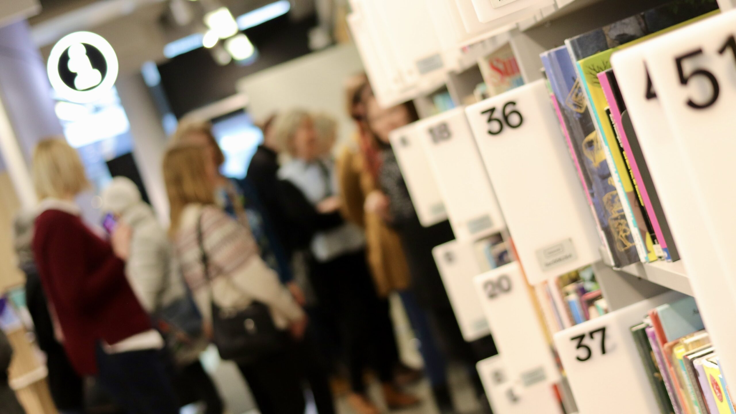 Numeroituja kirjahyllyjä etualalla, henkilöitä kirjastotilassa taustalla.