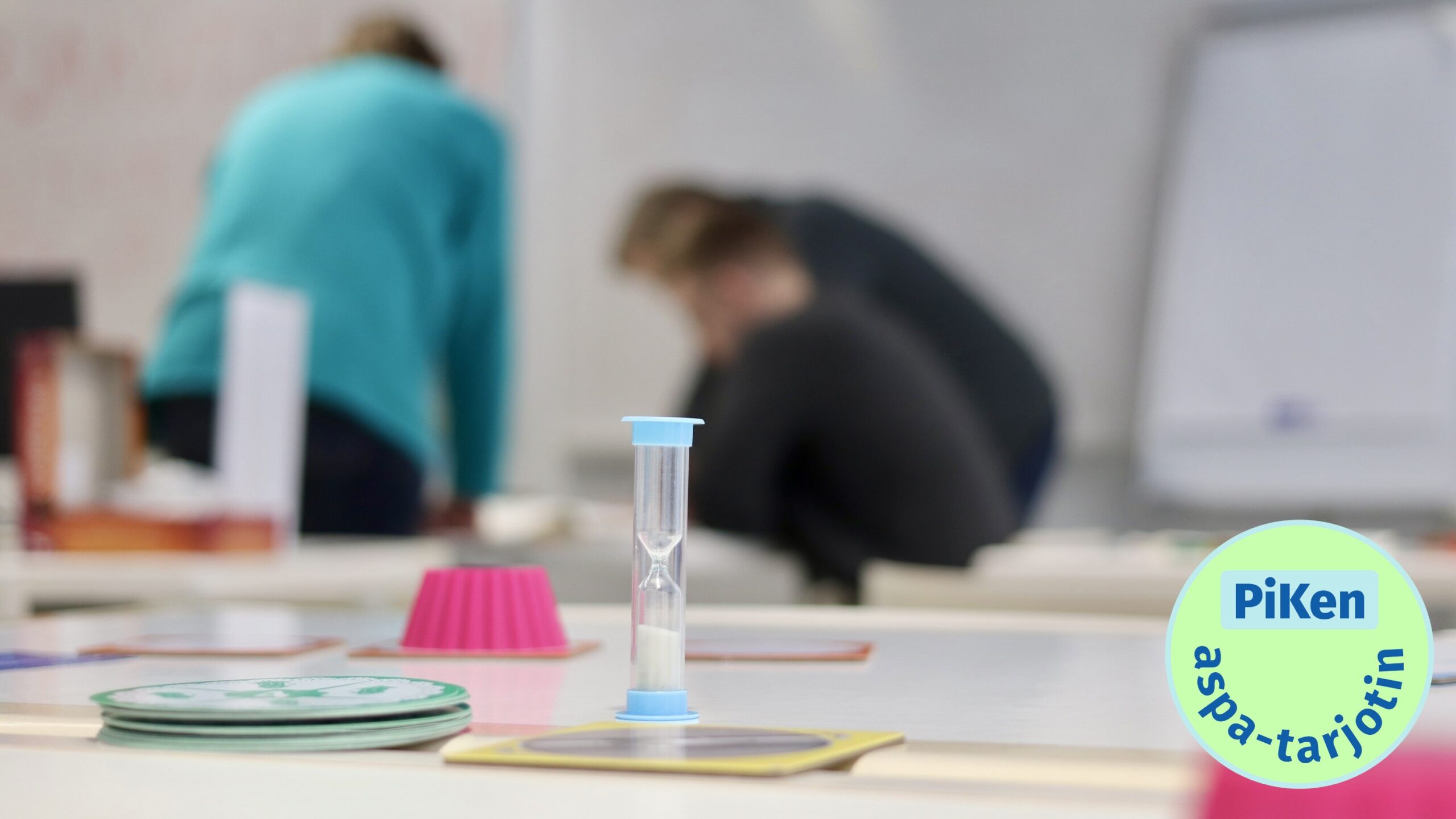 Etualalla tiimalasi ja pelimerkkejä. Takana muutama henkilö selin.