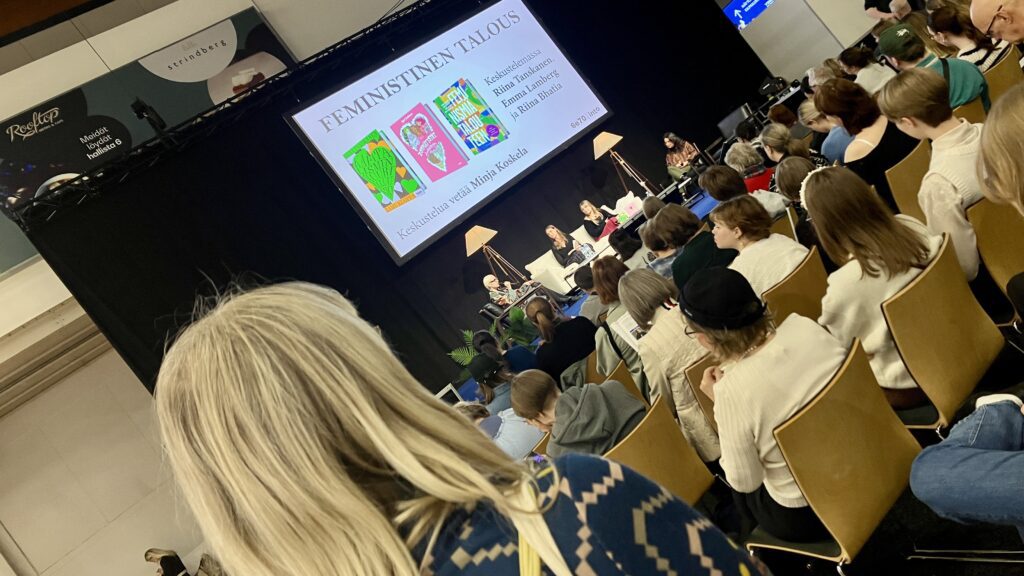 Ihmisiä selin kameraan seuraamassa lavan tapahtumaa aiheesta Feministinen talous.