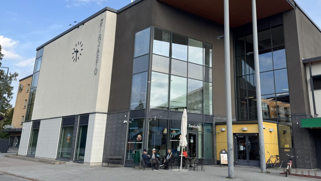 Kangasalan pääkirjaston sisäänkäynti poutaisena syyspäivänä. Kirjaston ulkopuolella istuu joukko ihmisiä terassituoleilla.