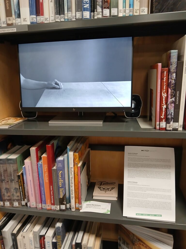 Tilaa elävöittävä videotaideteos Puijonlaakson lähikirjaston kirjahyllyssä. Pysäytyskuvassa teoksessa ihmisen käsi lepää pöydällä.