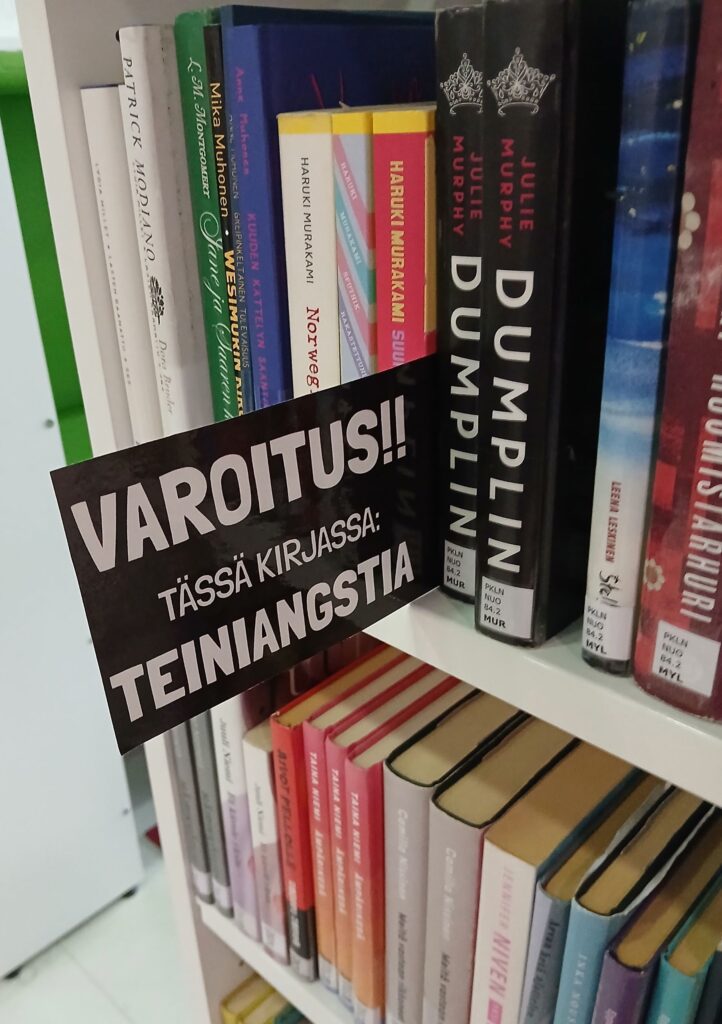Lähikuva kirjahyllystä, josta sojottaa kyltti "Varoitus!! Tässä kirjassa: teiniangstia".