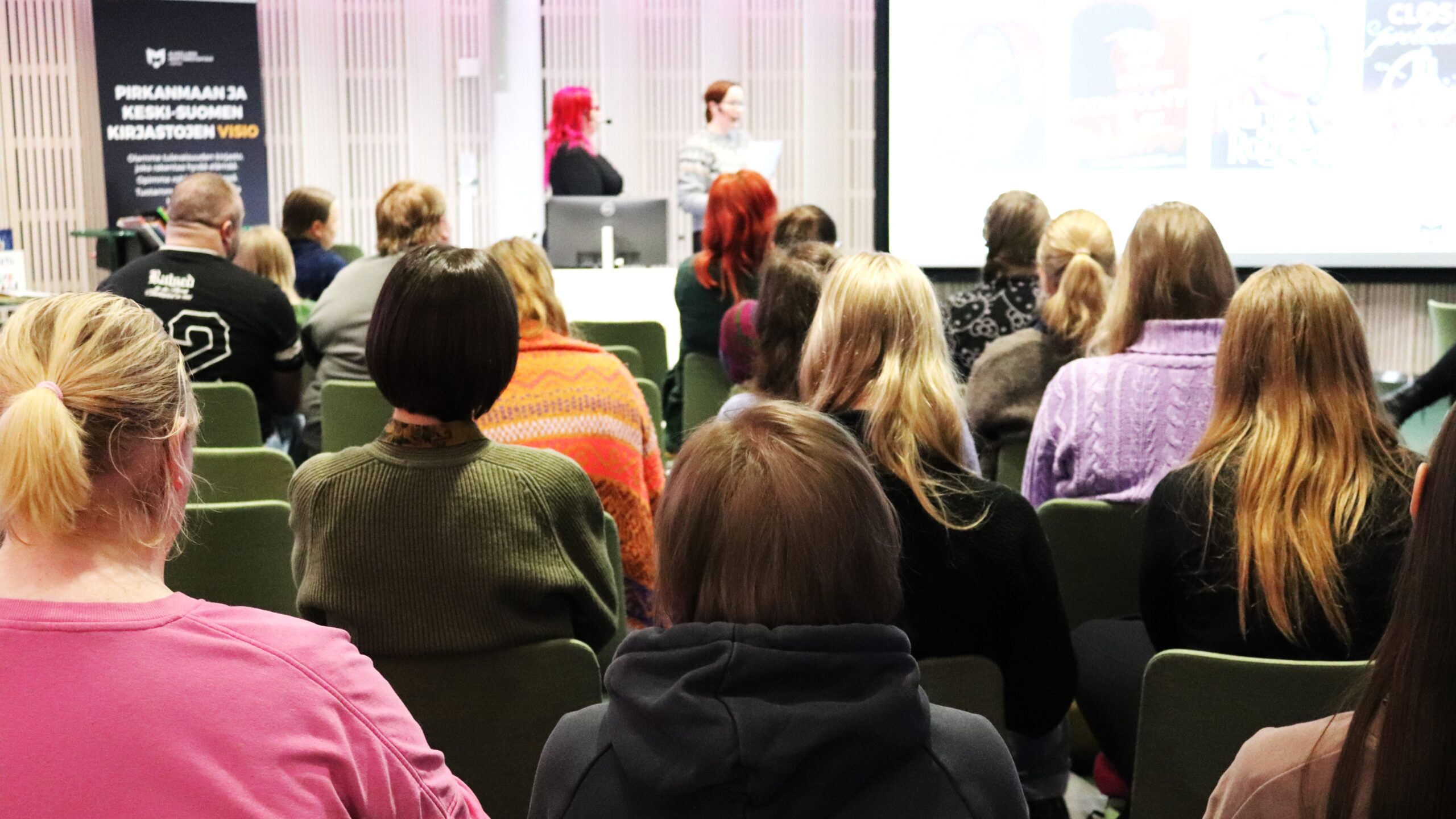 Ihmisiä selkä kameraan päin istumassa luentosalissa. Kaksi esiintyjää näkyy sumeina taustalla.