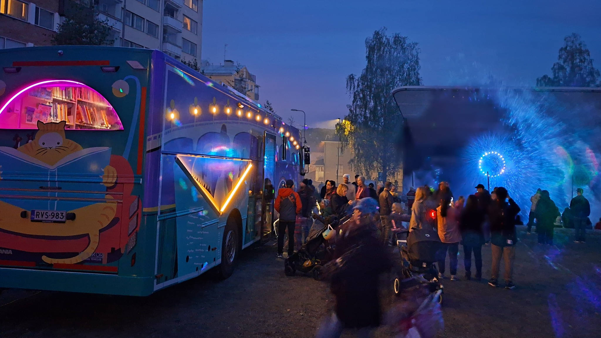 Jyväskylän kirjastoauto Wäinö kuvattuna Valon kaupunki -tapahtumassa vuonna 2025. Kirjastoauton valot loistavat hämärässä illassa, ympärillä kosolti kaupunkilaisia tutustumassa Wäinöön.