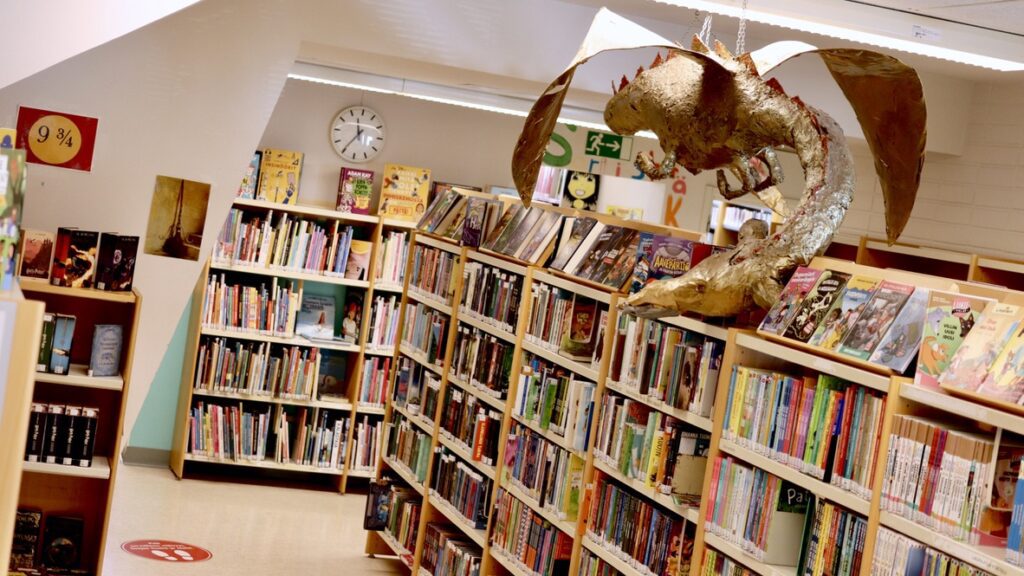 Paperimassasta muovattu kultainen lohikäärme liitelee Oriveden kirjaston kirjahyllyjen päällä.