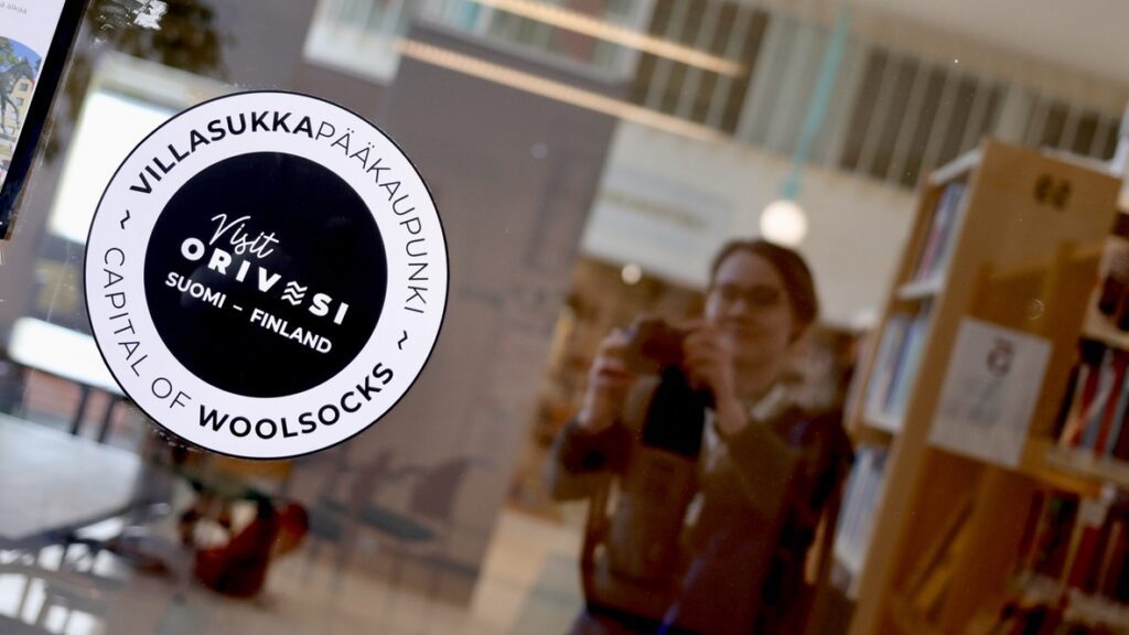 Orivesi - villasukkapääkaupunki -logo, taustalla valokuvaa ottava koordinaattori lasipinnan heijastuksena.