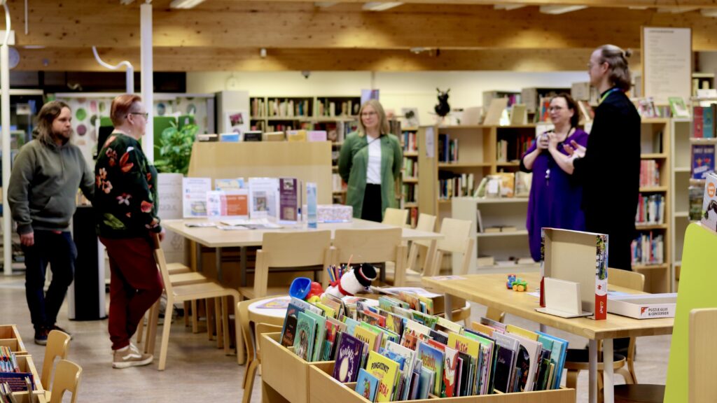 Petäjäveden kirjaston kirjastosali. Etualalla matalia kuvakirjahyllyjä. Taustalla henkilökuntaa ja AKE-koordinaattoreita keskustelemassa.