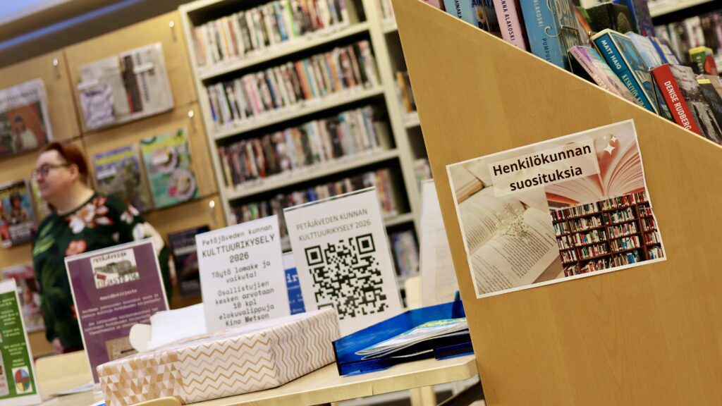 Petäjäveden kirjaston vaaleita puisia kirjahyllyjä ja pöytä, jossa Kulttuurikysely 2026 -juliste.