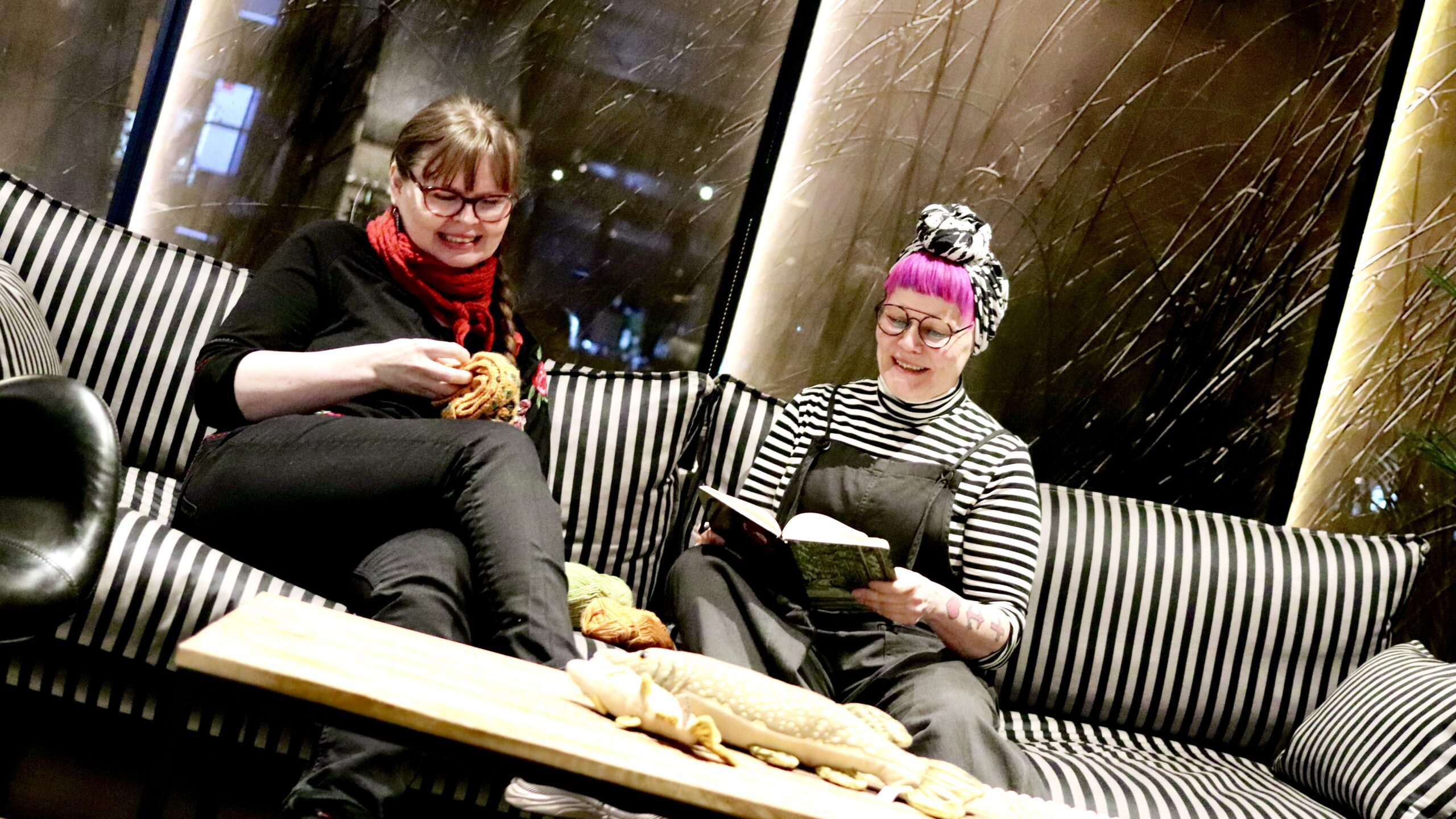 Kaksi henkilöä istumassa raidallisella sohvalla hymyillen, kirja ja virkkuutyö käsissään.