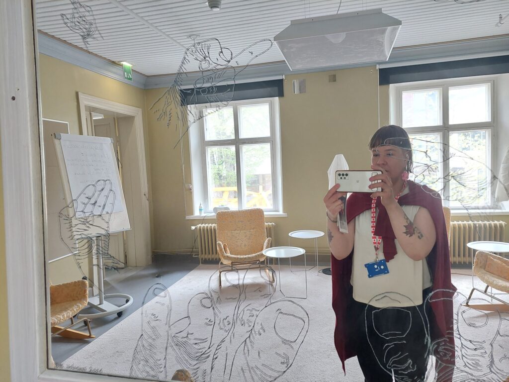 Kirjastonhoitaja valokuvaamassa itseään peilin kautta valoisassa huoneessa. Peilissä on ihmisen käsiä ja jalkoja kuvaavia ääriviivapiirustuksia, taustalla huoneessa fläppitaulu ja pehmeältä näyttävä matto.