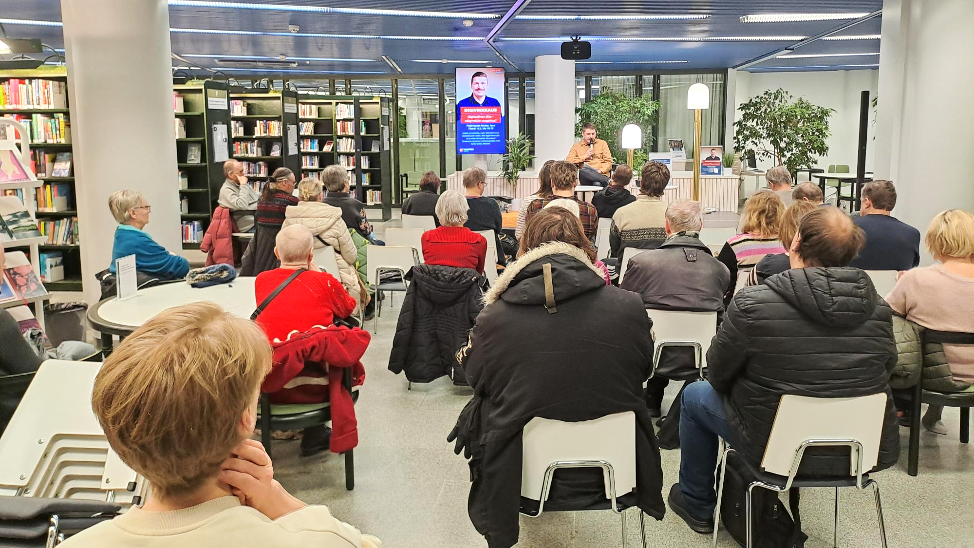 Ihmisiä istumassa tuoleilla kirjastotilassa selät kameraan päin. Mies puhumassa heidän edessään mikrofoniin.