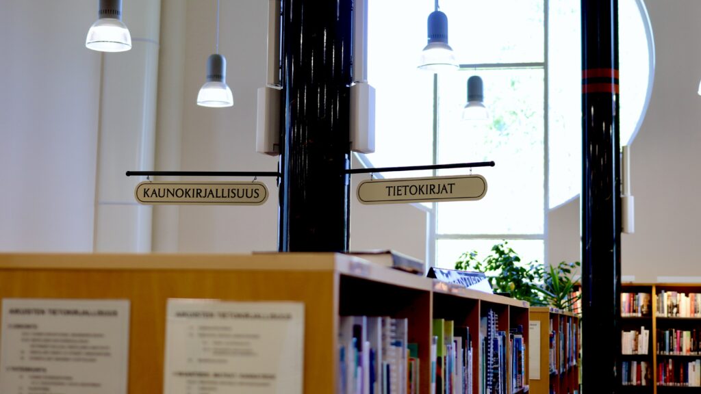 Kirjahyllyjä ja opasteita Pälkäneen kirjastossa, taustalla valoisa ikkuna.
