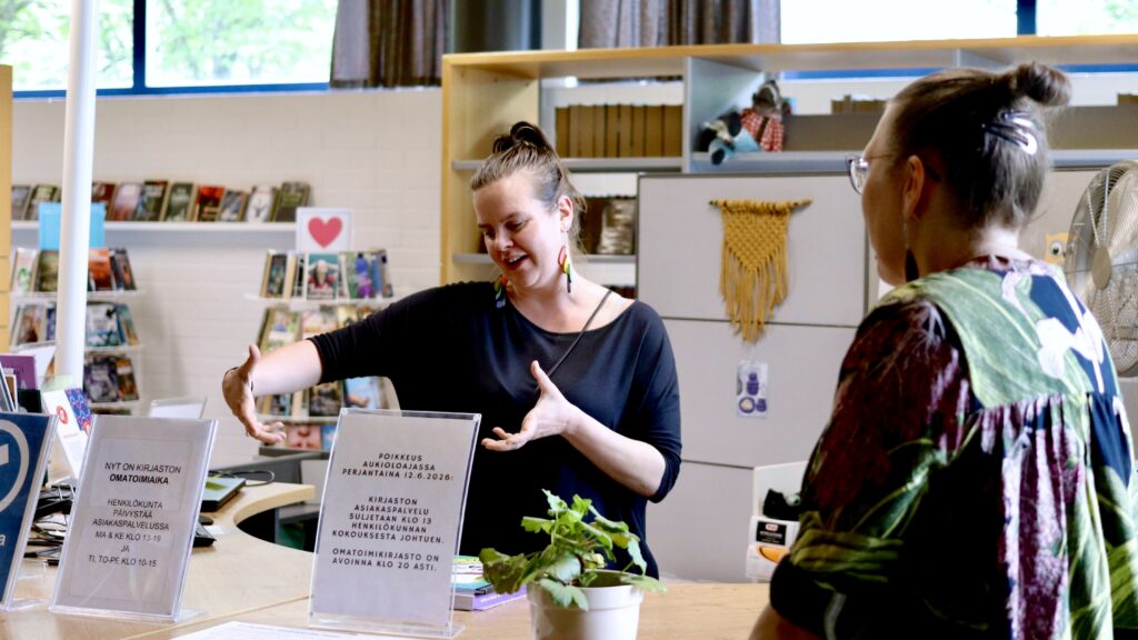 Juupajoen kirjaston henkilökunnan edustaja elehtii puhuessaan käsillään kirjaston asiakaspalvelutiskin takana. Tiskillä kaksi kylttiä, joissa kerrotaan omatoimikirjaston toiminnasta ja kirjaston palveluajoista.