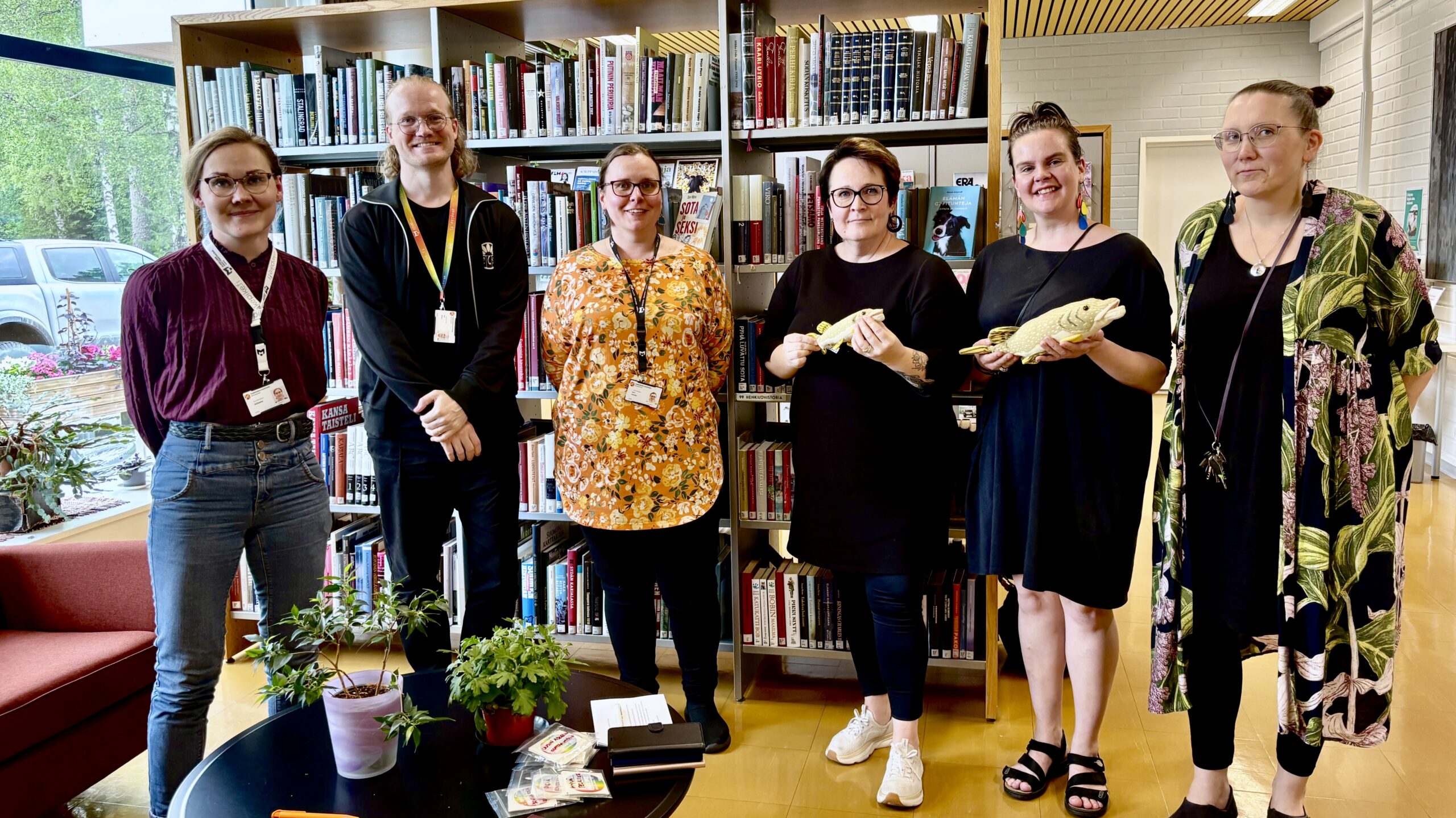 Hymyileviä kirjastoammattilaisia Juupajoen kirjastossa. Taustalla kirjahylly, ikkunasta näkyy täydessä lehdessä olevia koivuja.